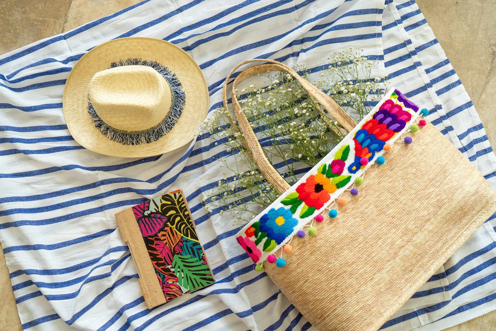 Woven Mexican tote bag with colorful embroidered flowers and pom-poms beside a straw hat on striped fabric